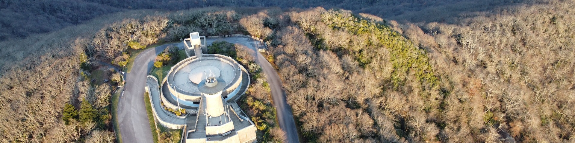 An aerial view of a circular building complex surrounded by a winding road, set against a backdrop of forested hills.
