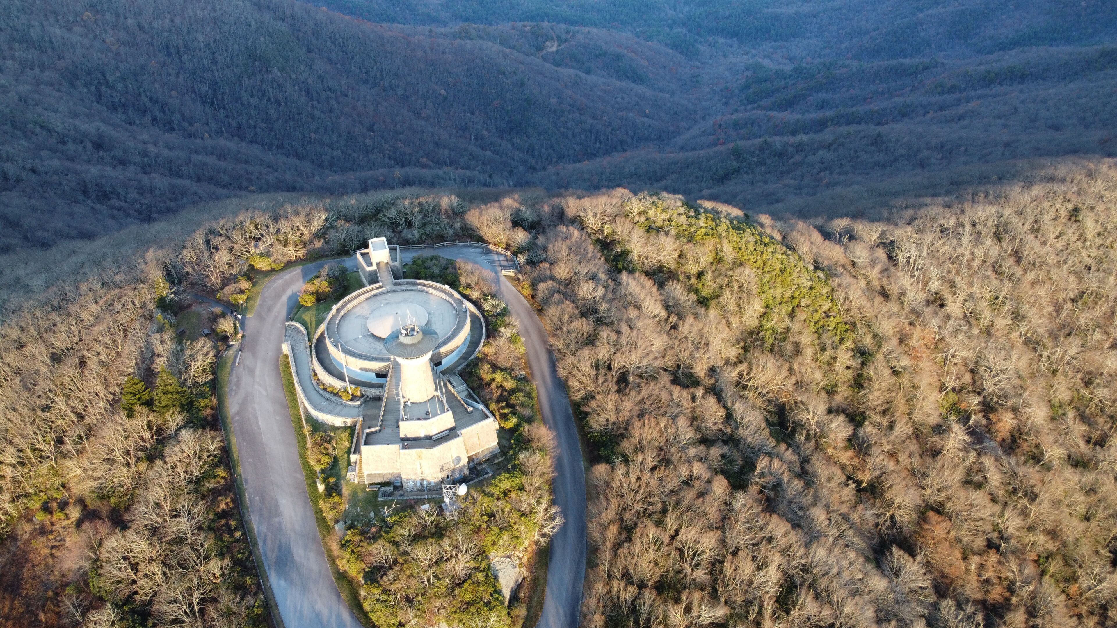 An aerial view of a circular building complex surrounded by a winding road, set against a backdrop of forested hills.