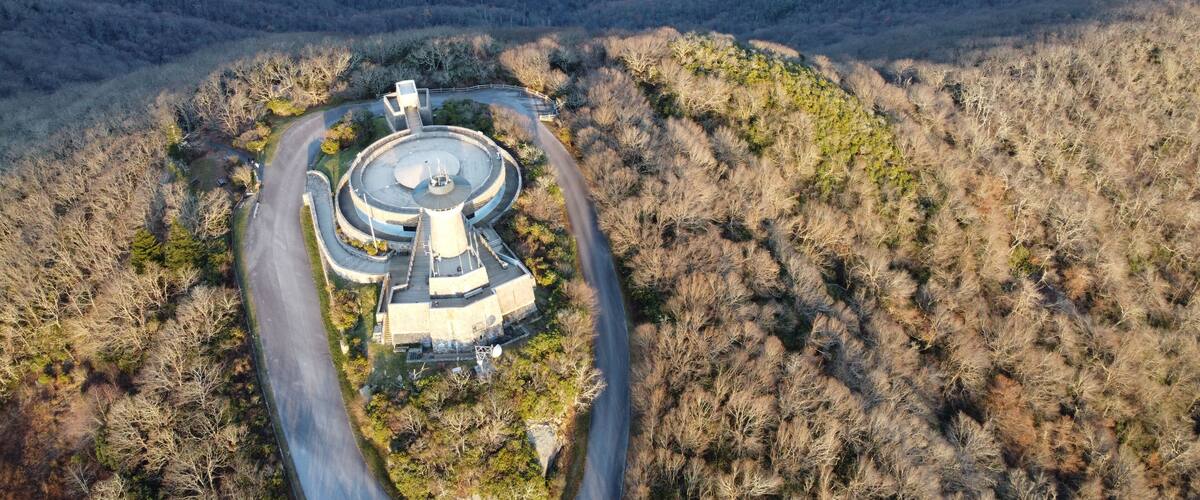 An aerial view of a circular building complex surrounded by a winding road, set against a backdrop of forested hills.