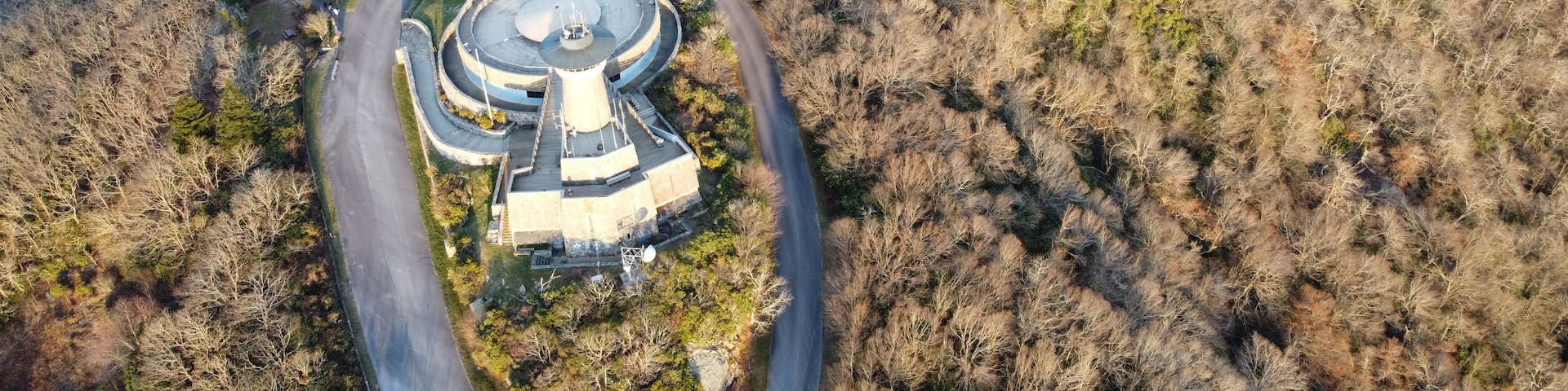 An aerial view of a circular building surrounded by a winding road, set in a hilly forested area.