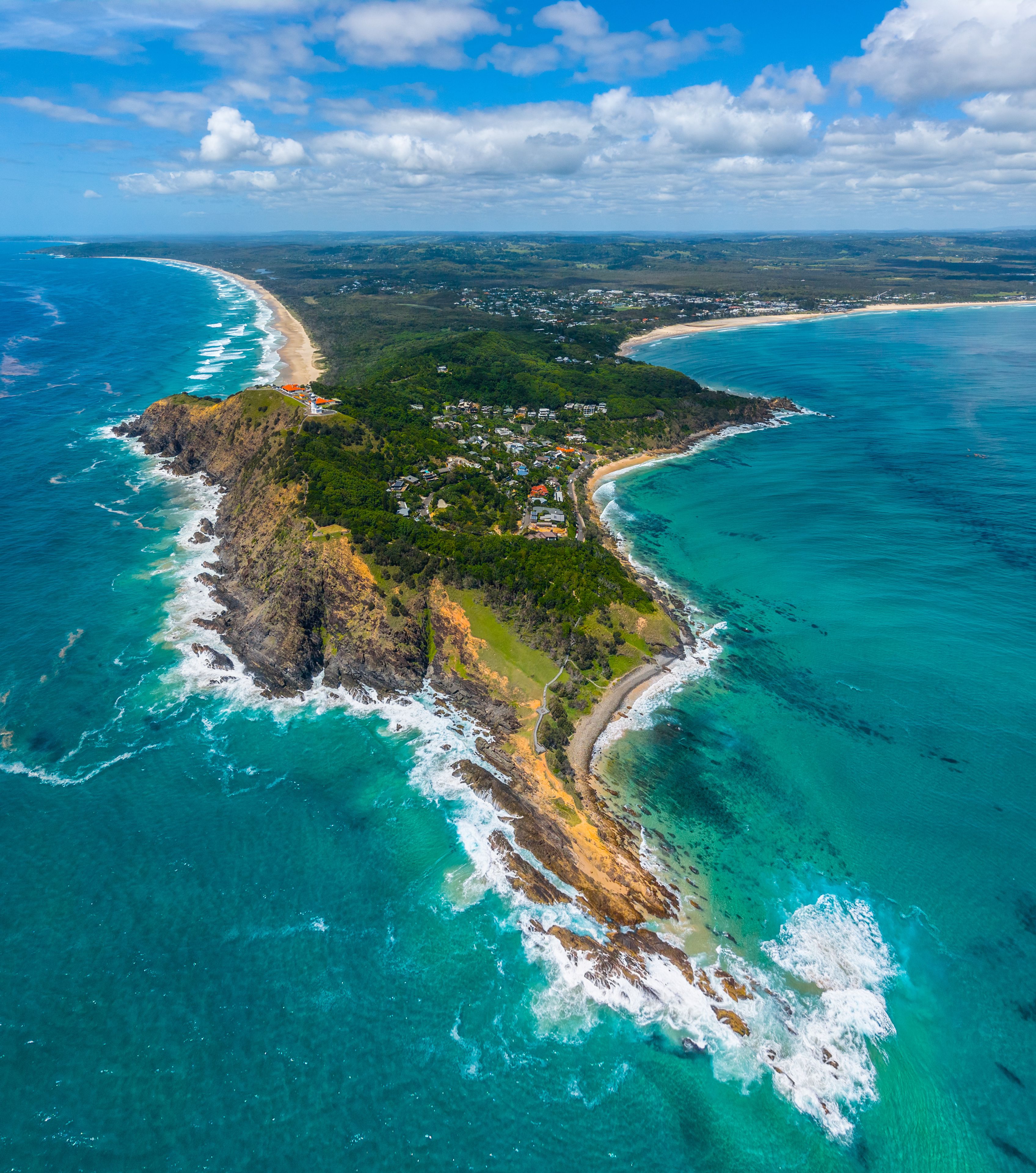 An aerial view of a coastal area with a curving beach, dense green vegetation, and clear turquoise waters.