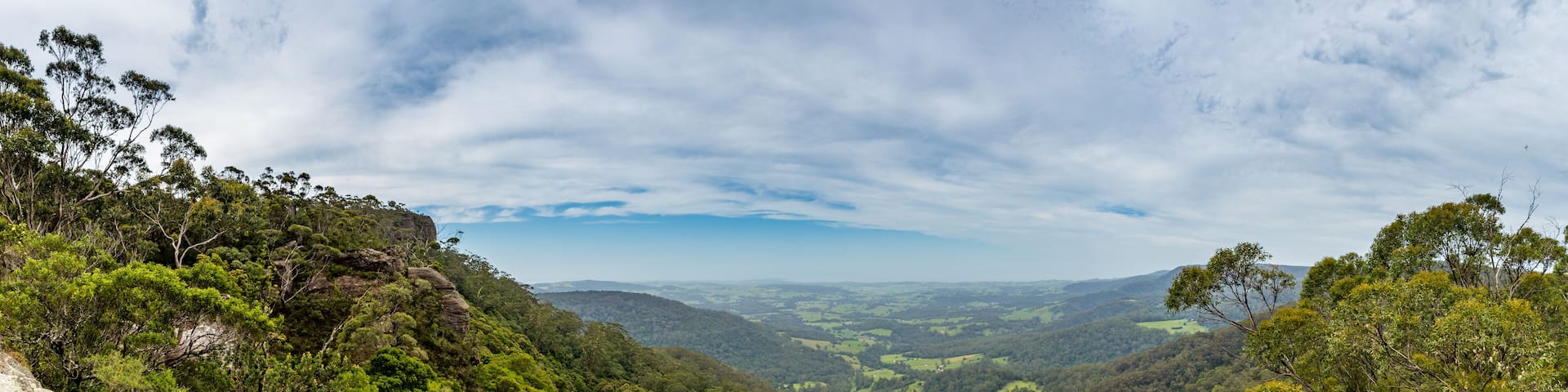 A panoramic view of a lush green valley with rolling hills and trees from a clifftop in Jamberoo.