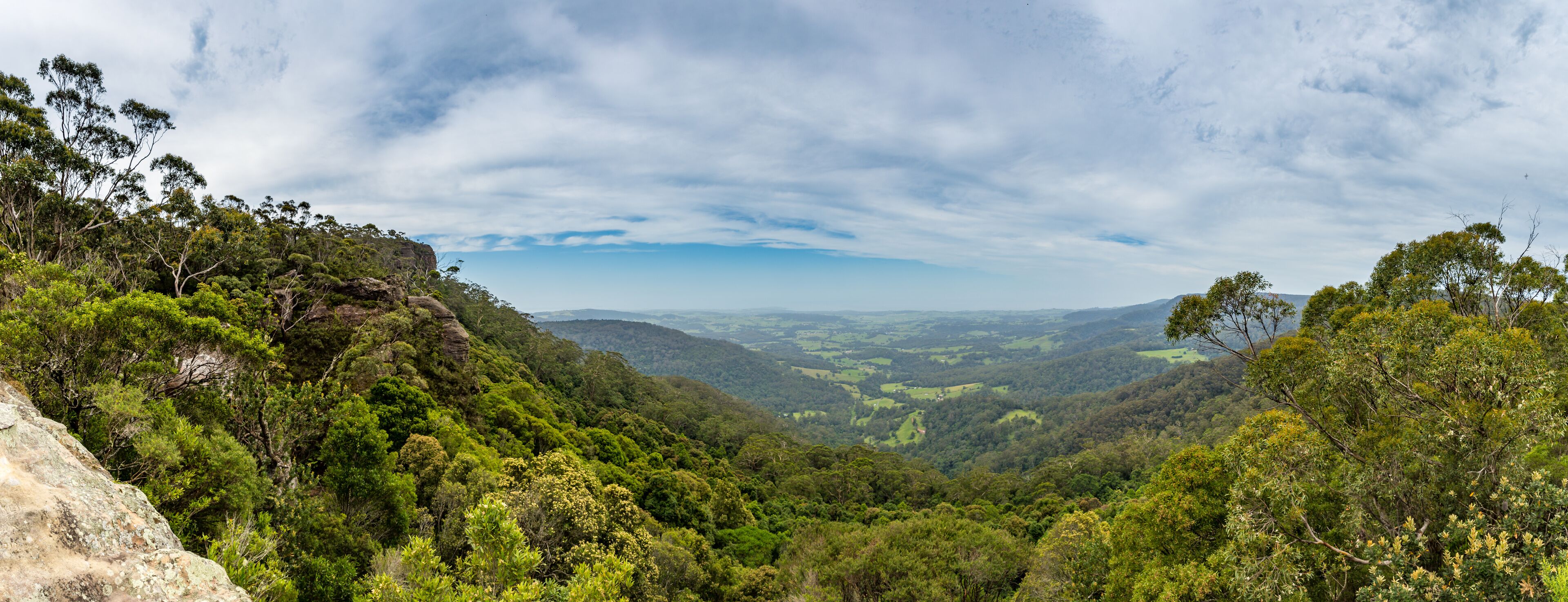 A lush valley with dense green foliage, rocky outcrops, and a distant mountain range under a partly cloudy sky.