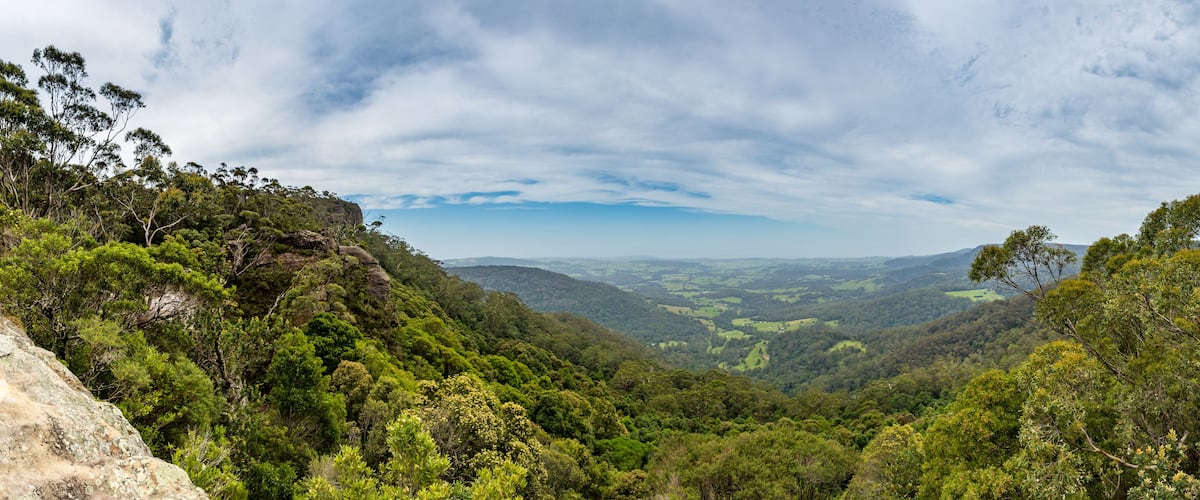 A lush valley with dense green foliage, rocky outcrops, and a distant mountain range under a partly cloudy sky.