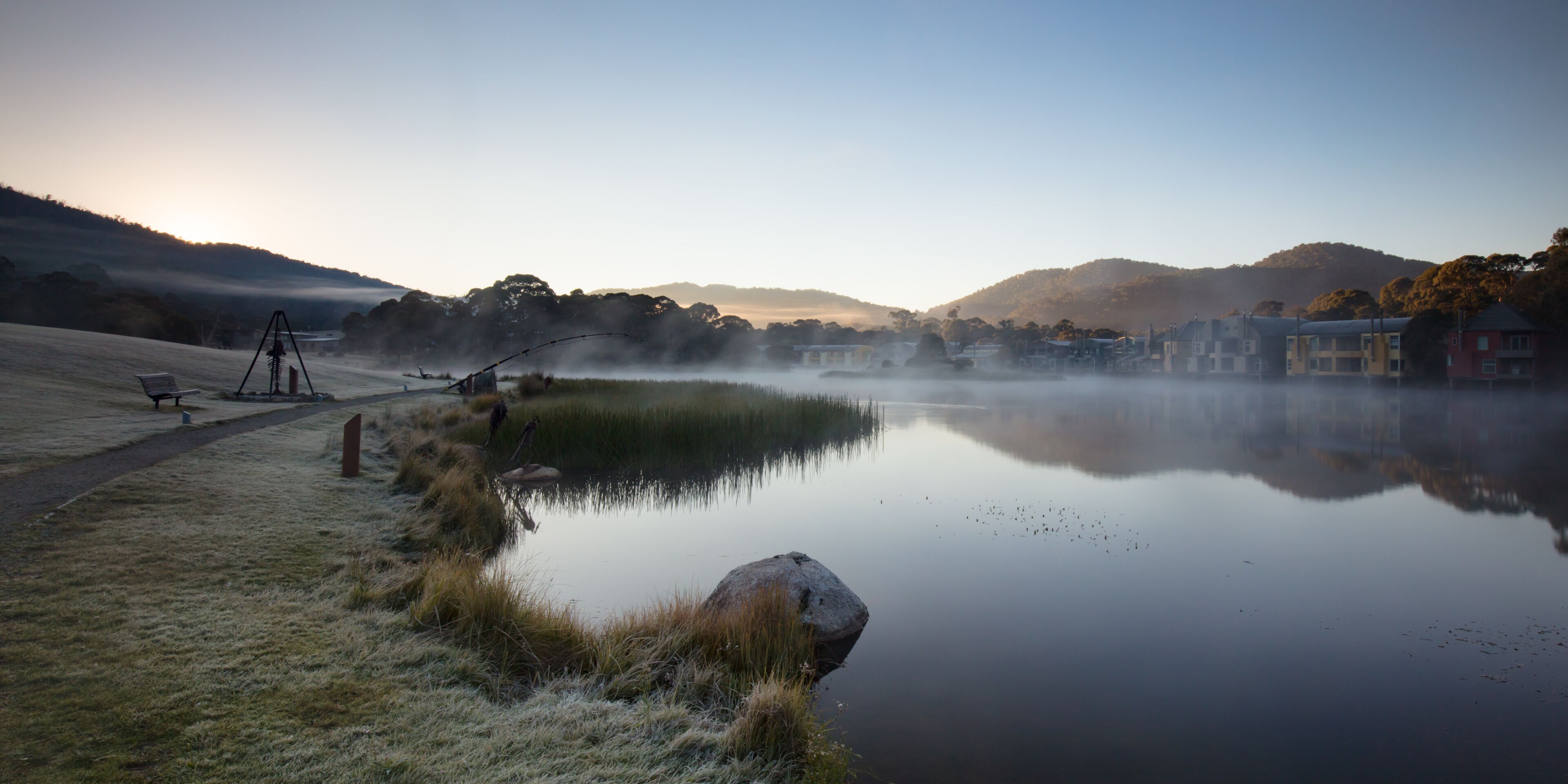A calm lake with a tripod, grass, a rock, and buildings in the background.