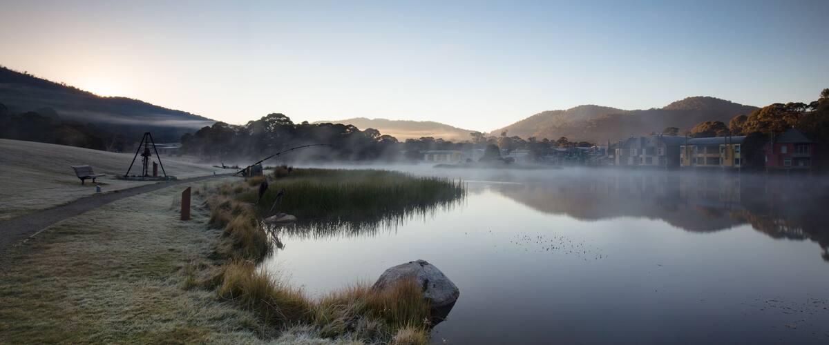 A calm lake with a tripod, grass, a rock, and buildings in the background.