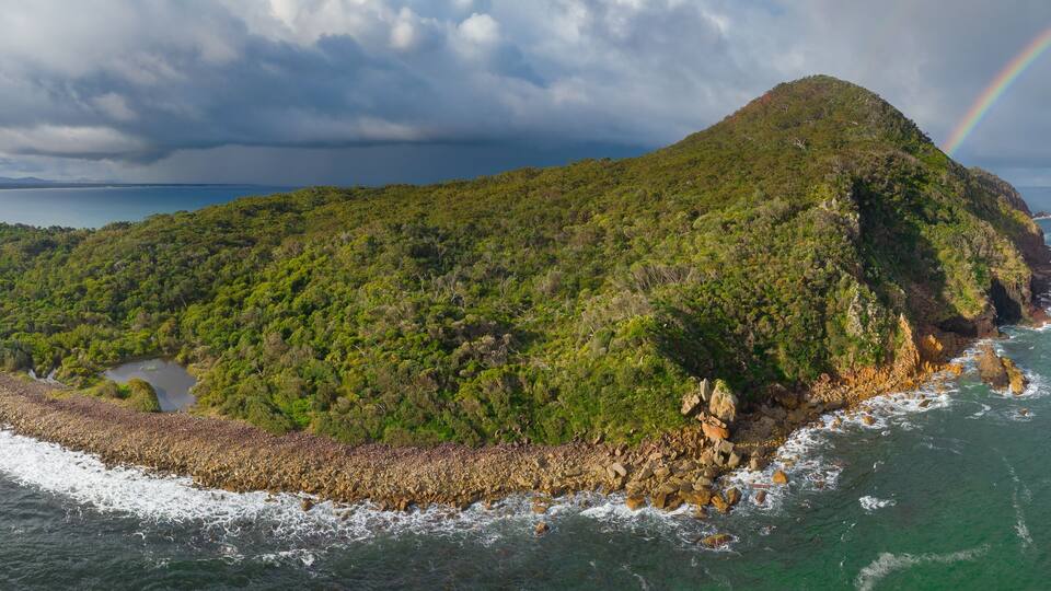 A lush green island surrounded by ocean waves, with a visible rainbow and a distant shoreline.