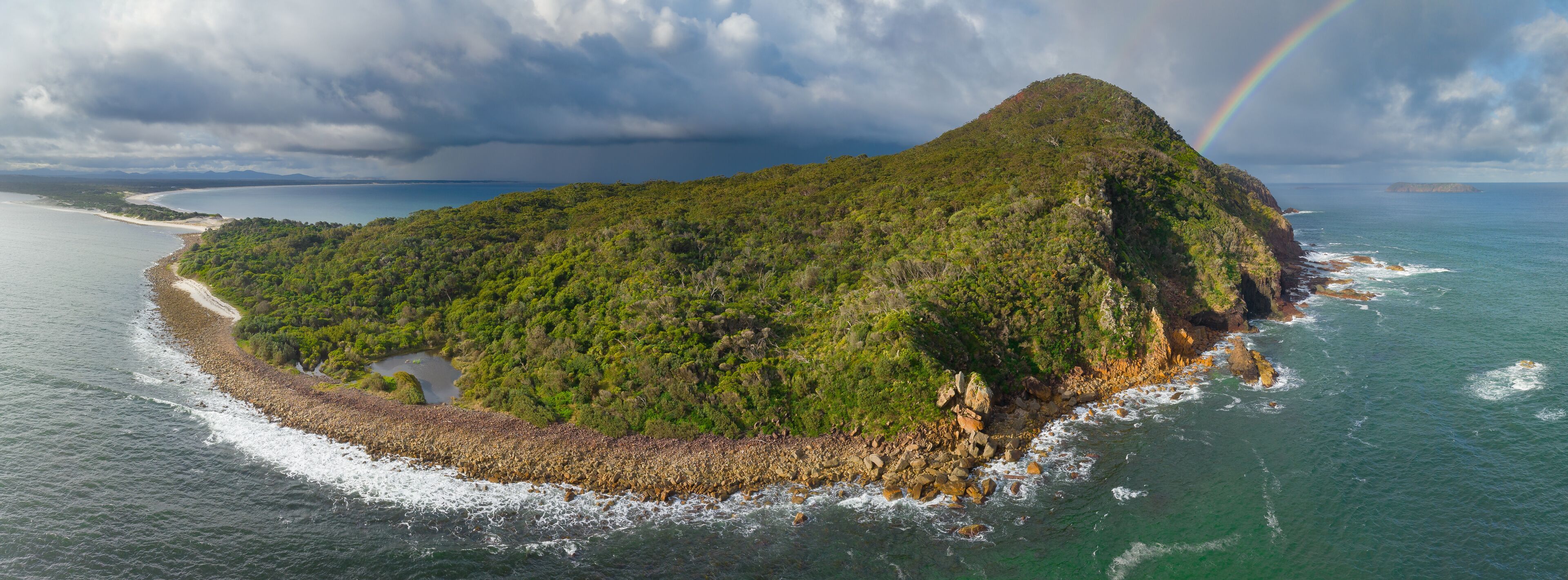 A lush green island surrounded by ocean waves, with a visible rainbow and a distant shoreline.
