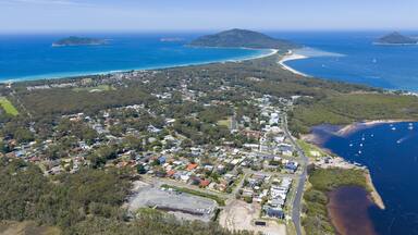 An aerial view of a coastal town with a beach, clear blue waters, and a mountainous island in the distance.