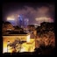 Paisatge urbà nocturn amb edificis il·luminats i primer pla d'arbres.