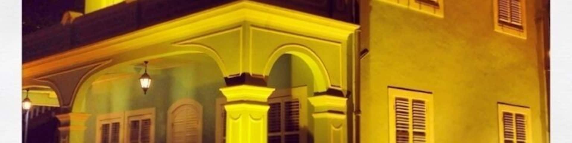 Un edificio de dos pisos con balcón y ventanas arqueadas, iluminado por luces amarillas.