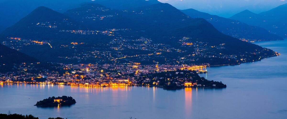 A coastal city at dusk with illuminated buildings and mountains in the background.