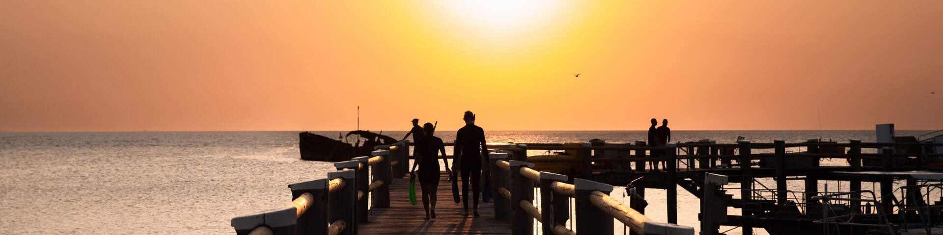 Un muelle de madera que se adentra en el océano al atardecer, con siluetas de personas caminando.