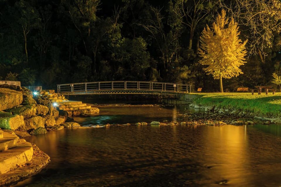 A stone bridge over a calm river at night, surrounded by trees and illuminated by artificial lights.
