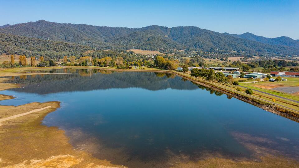 A tranquil lake with a clear reflection, surrounded by a rural landscape with fields, trees, and distant mountains.