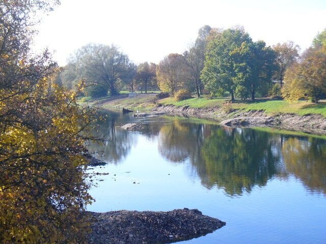 A calm river with trees reflecting on the water, surrounded by grassy banks and autumn foliage.