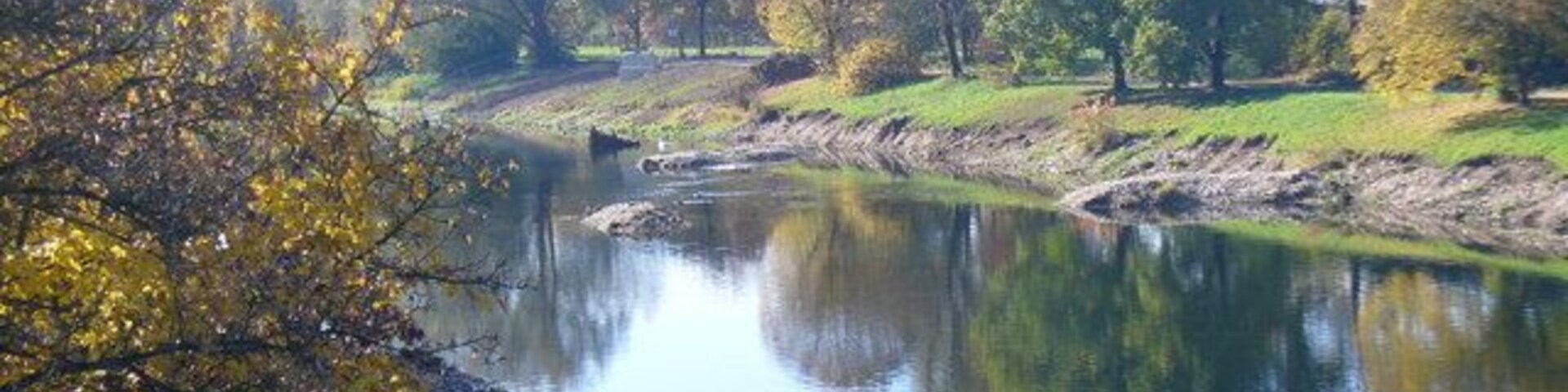 A calm river with trees reflecting on the water, surrounded by grassy banks and autumn foliage.