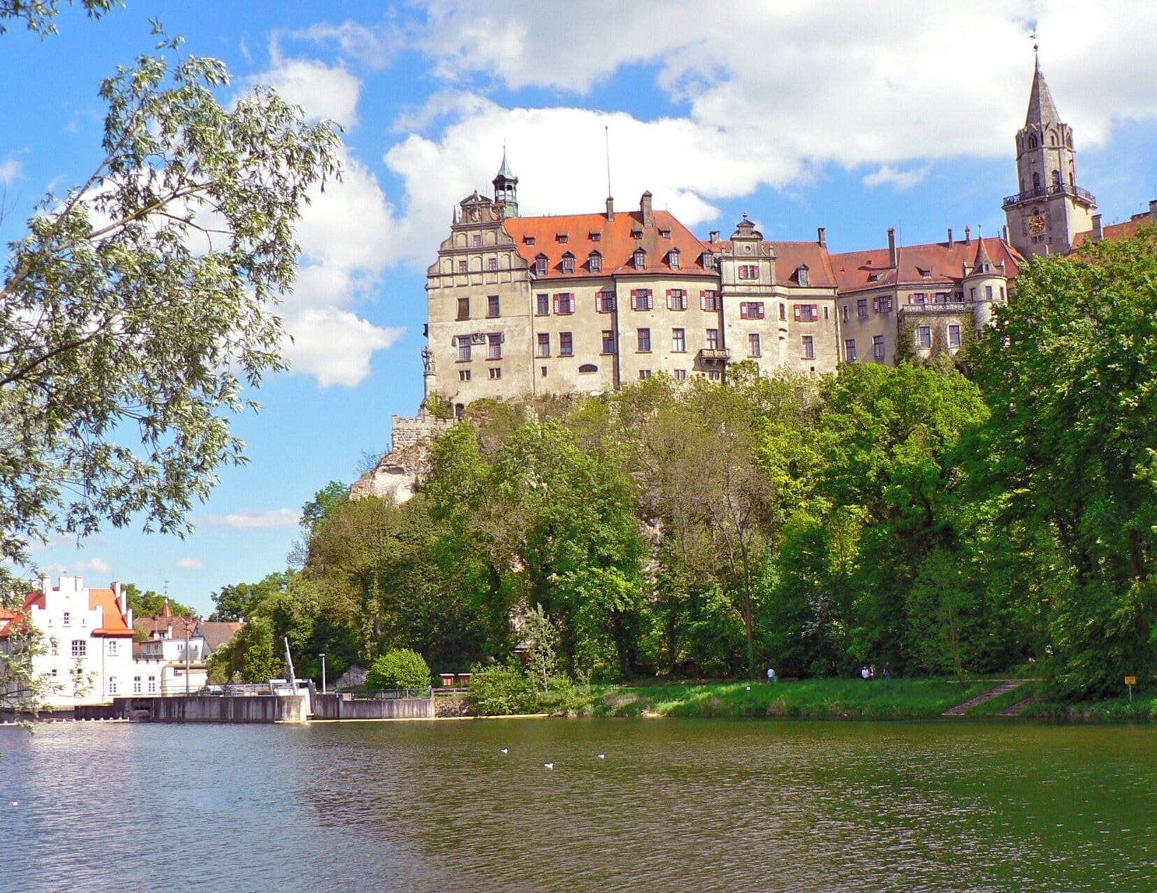 A historic castle on a hill overlooking a river, surrounded by lush greenery and a clear blue sky.