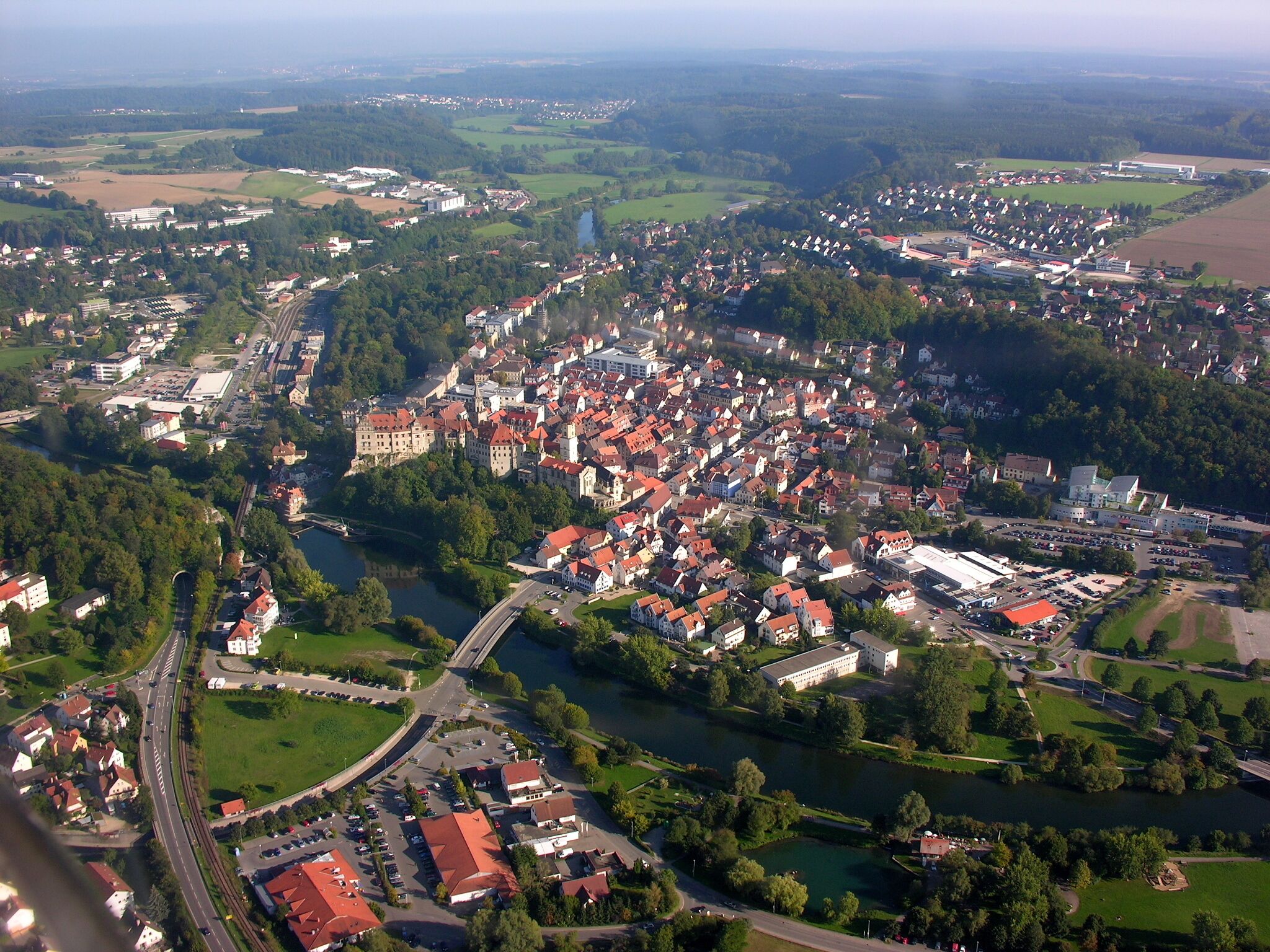 An aerial view of a town with a river, surrounded by greenery and featuring a castle-like structure.