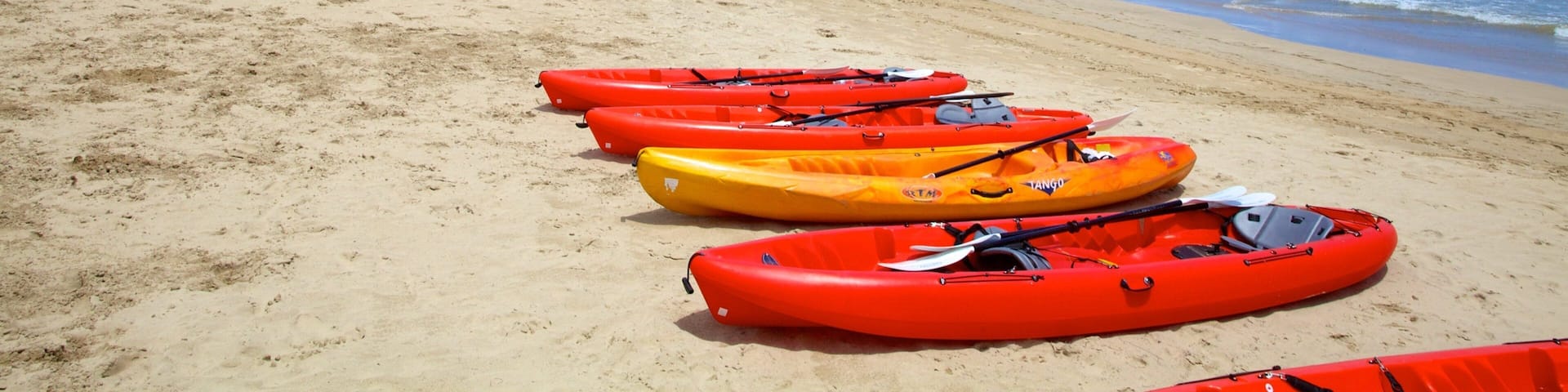 A beach with kayaks lined up on the sand and a kite surfer in the air.