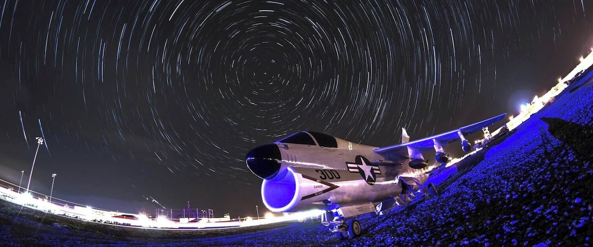 A night scene with a star trail effect, a military aircraft, and a crowd of people.