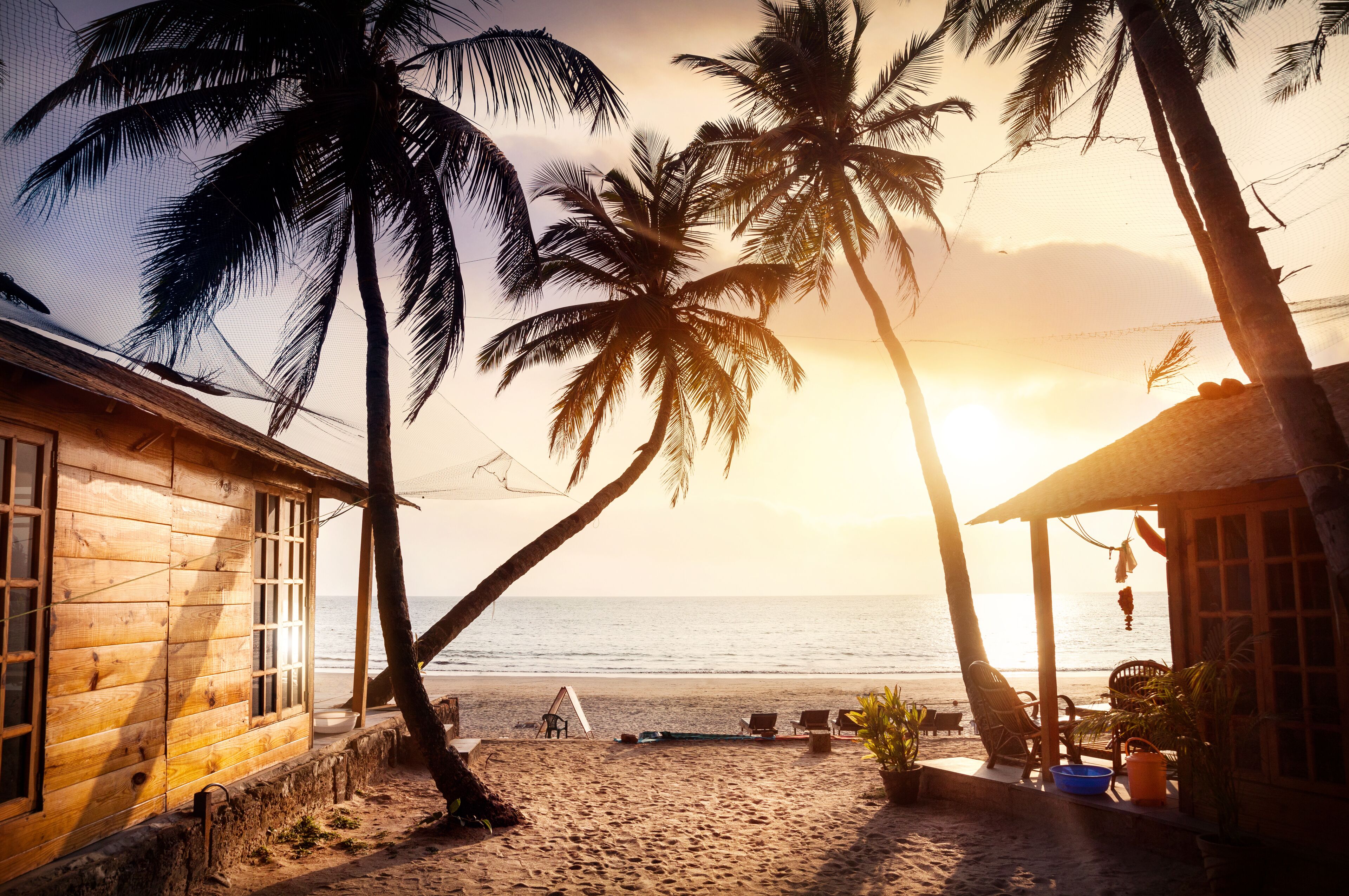 A wooden beach house with a view of the ocean at sunset, palm trees, and beach chairs.