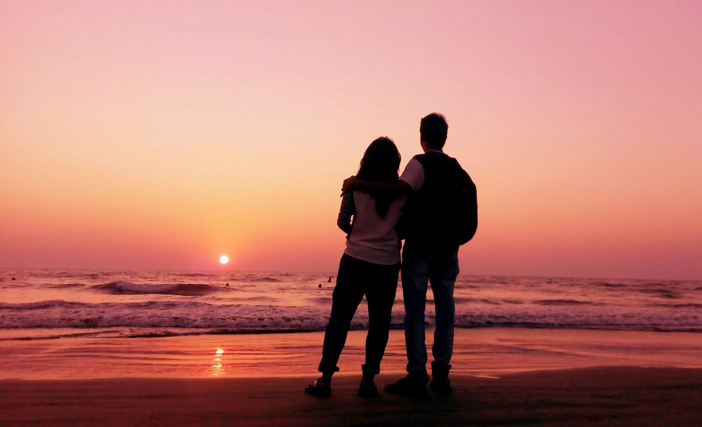 Two people standing on a beach at sunset with the sun low on the horizon.