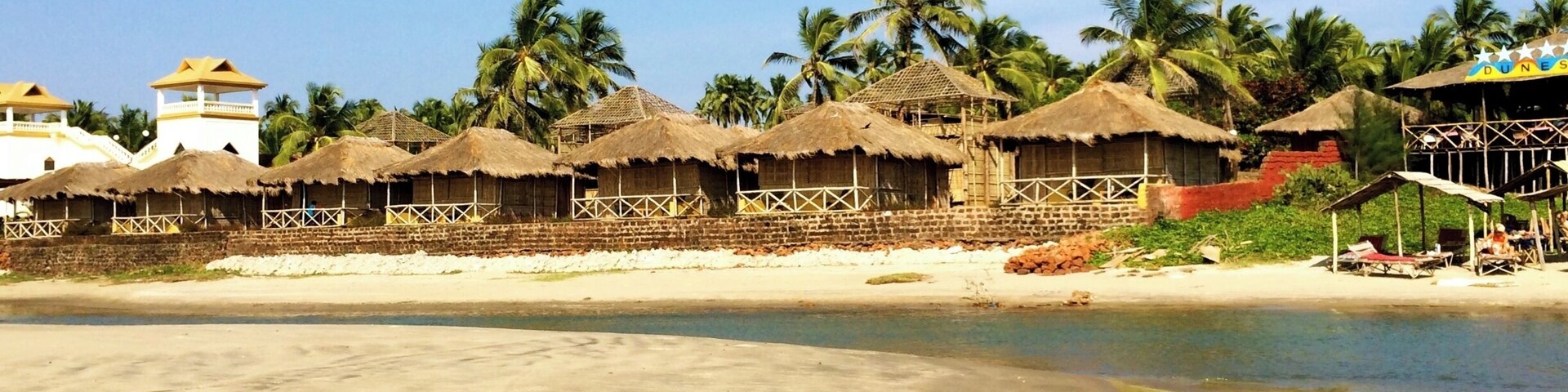Una playa con cabañas de techo de paja, palmeras y un edificio blanco con techo amarillo.