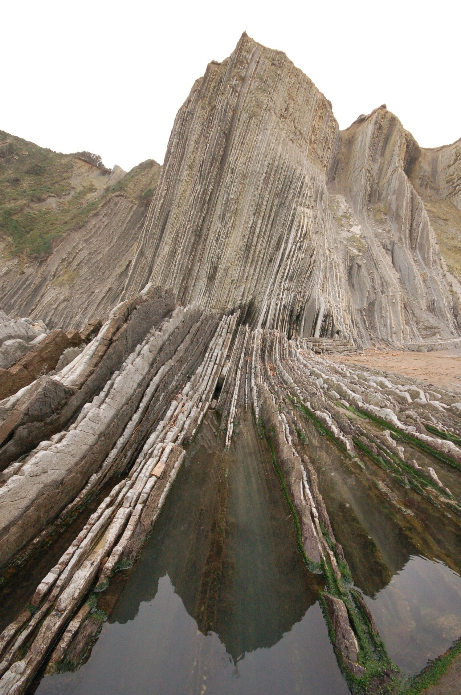 A large rock formation with steep, layered cliffs and a reflective pool at its base.