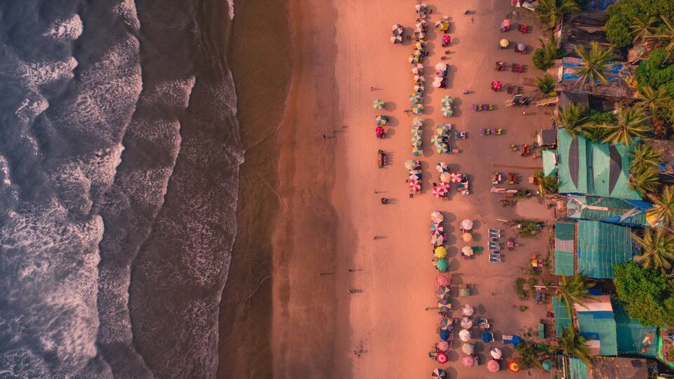 An aerial view of a beach with waves, umbrellas, and palm trees.