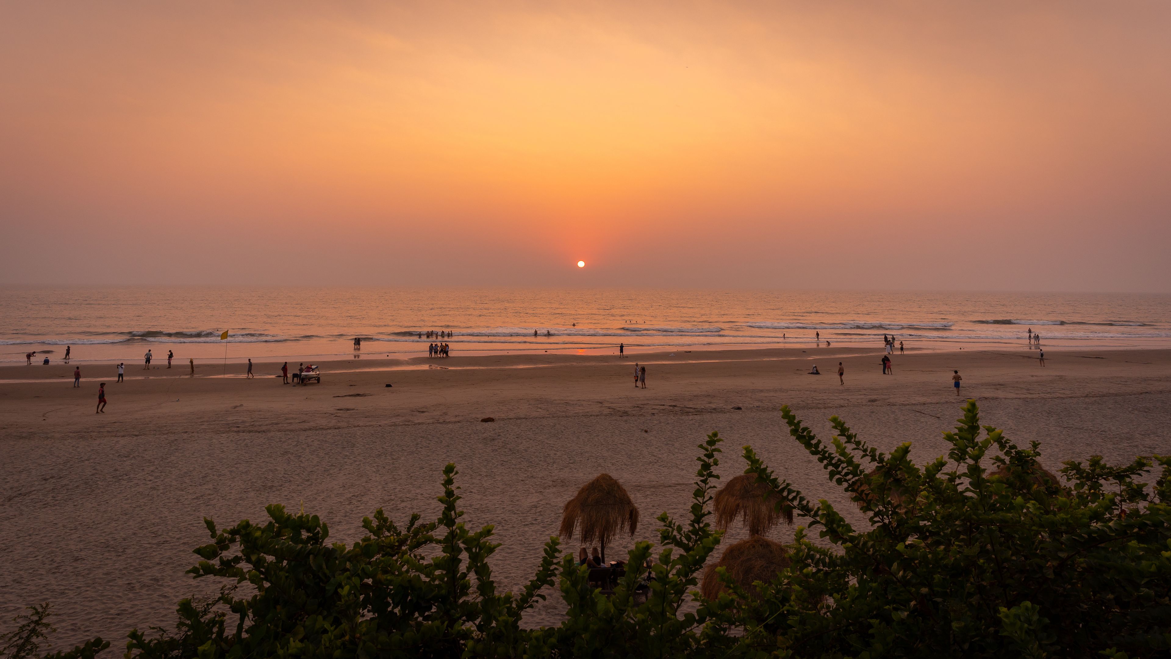 Uma praia ao pôr do sol, com pessoas caminhando e brincando, e guarda-sóis de palha em primeiro plano.