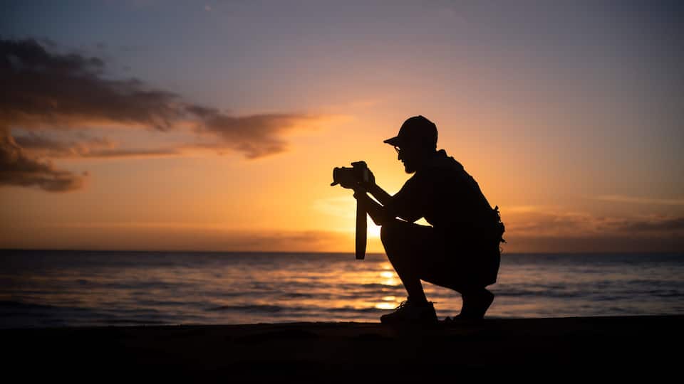 A silhouette of a person taking a photo at sunset by the ocean.