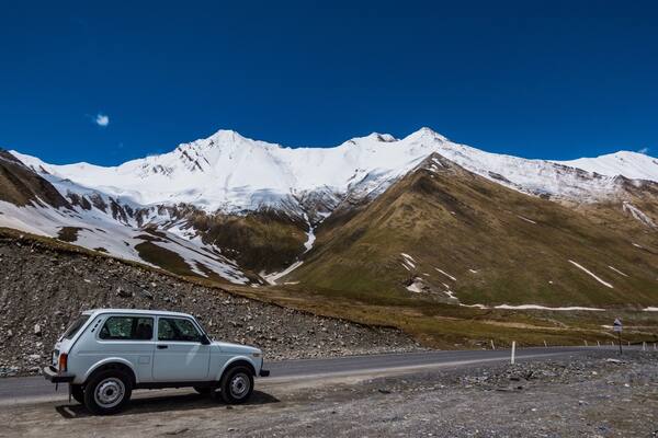 A white car on a mountain road with snow-capped peaks in the background.