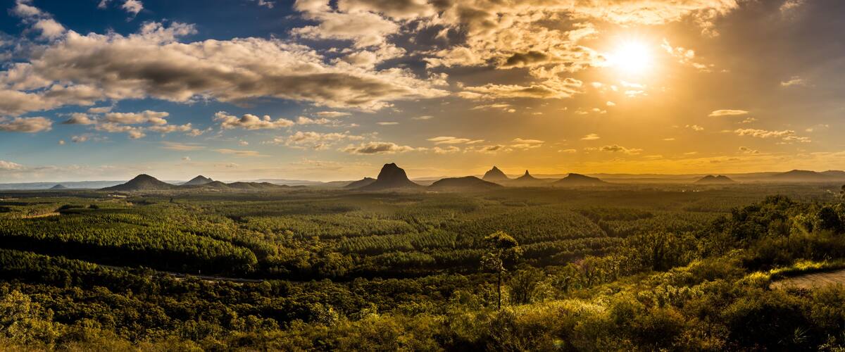 A landscape with a prominent mountain peak, dense forest, and a clear sky with the sun setting or rising.