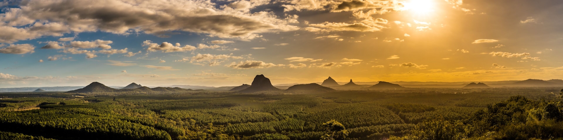 A landscape with a prominent mountain peak, dense forest, and a clear sky with the sun setting or rising.