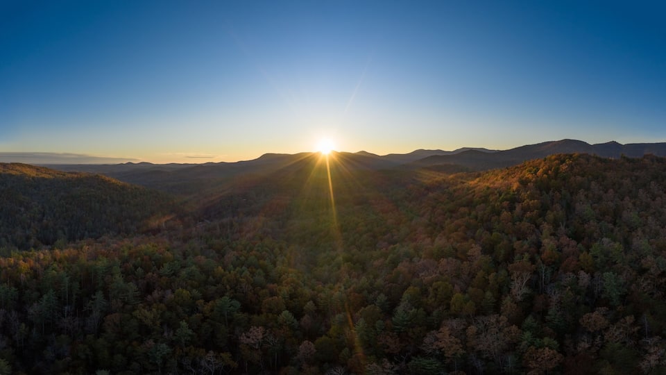 A mountainous landscape with a sunrise or sunset, casting rays of light through the trees.