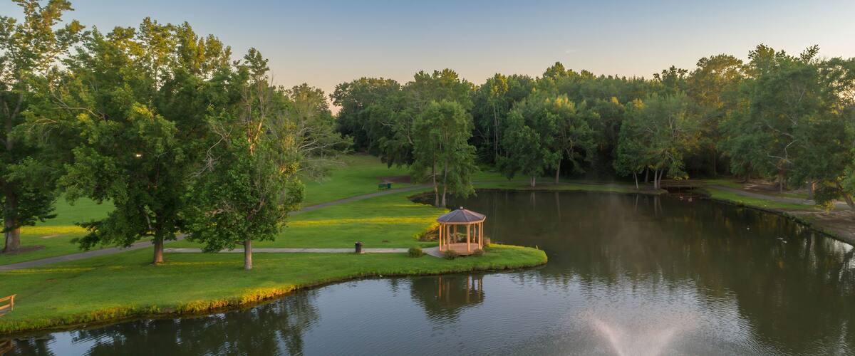 Aerial view of Fairview Park, Dublin, Georgia, USA, featuring a gazebo on a peninsula, a fountain in the lake, and lush green trees. The scene captures the tranquility of nature.