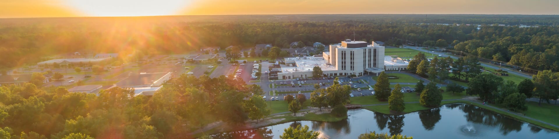 Aerial view of a hospital complex Fairview Park, Dublin, Georgia, USA. The building is surrounded by trees, a pond, and parking lots. The sun is setting in the distance.