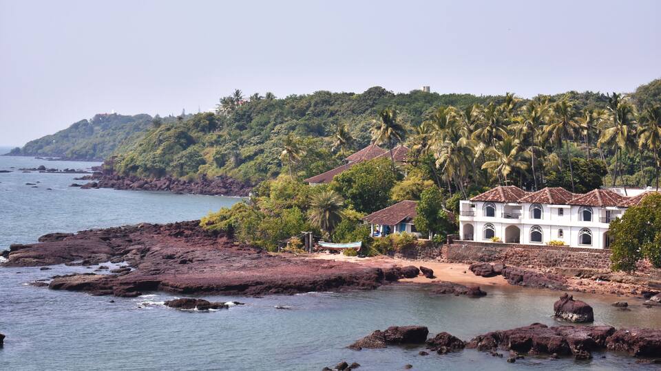 A coastal landscape with rocky shores, a white building with a red roof, and dense palm trees on a hillside.