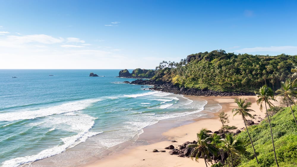 A tropical beach with clear blue waters, sandy shore, and lush green hills.