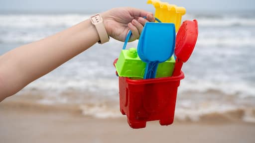 Woman holding up a colorful sand castle making kit with bucket, molds, shovel against the waves on baga, morjim, vagator, paolem beach in goa a popular tourist spot