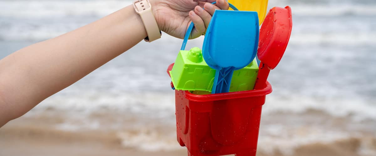 A hand holding a beach toy bucket with a blue shovel and a yellow toy.