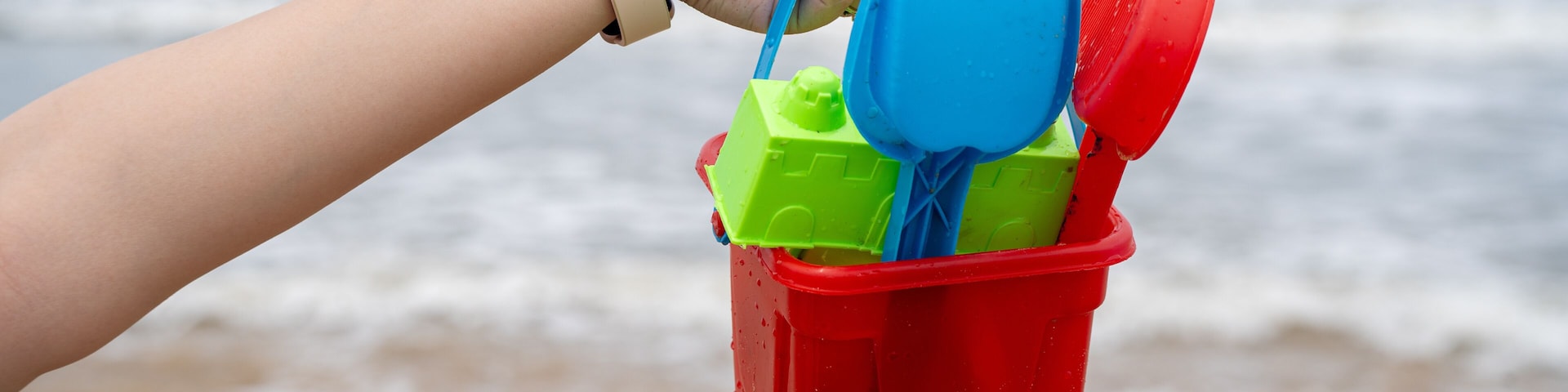 A hand holding a beach toy bucket with a blue shovel and a yellow toy.