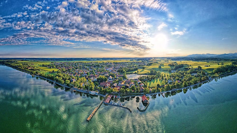 A panoramic view of a lakeside town with a bridge, boats, and surrounding greenery.