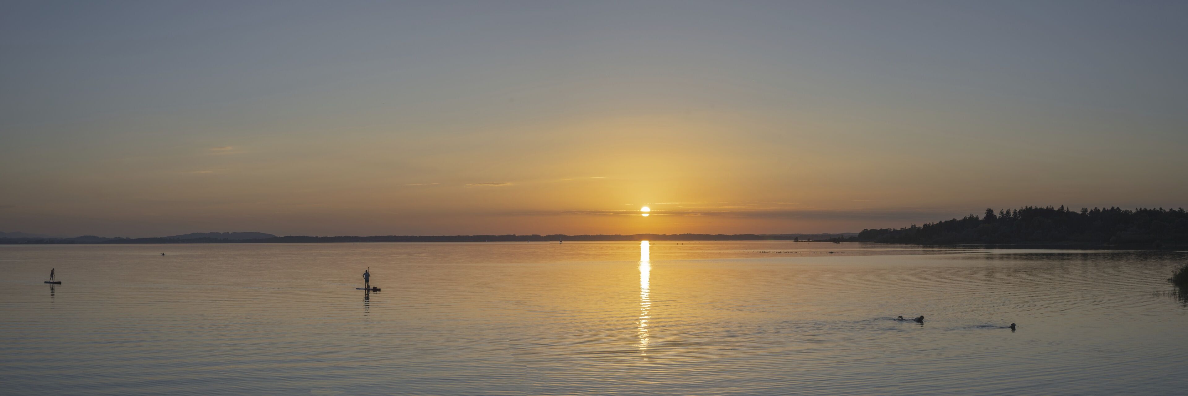 A calm lake at sunset with a visible sun reflection, a distant shoreline, and a few people paddling.