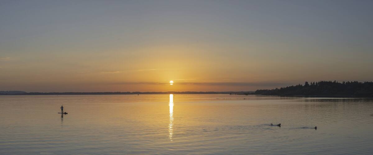A calm lake at sunset with a visible sun reflection, a distant shoreline, and a few people paddling.
