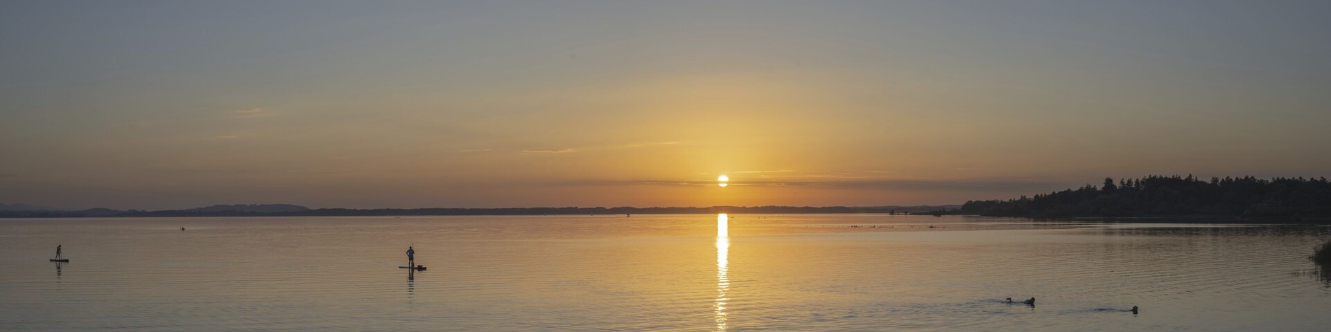 A calm lake at sunset with a visible sun reflection, a distant shoreline, and a few people paddling.