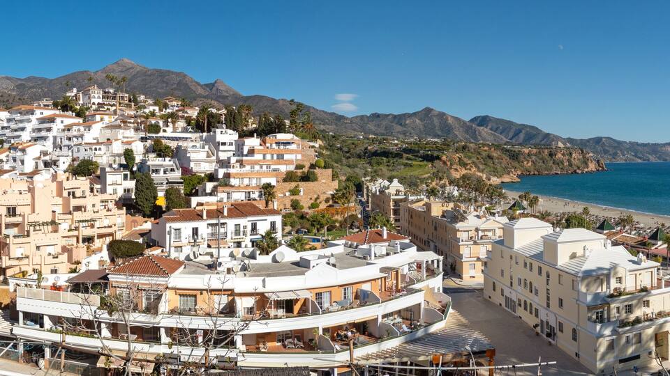 A coastal town with white buildings, a beach, and mountains in the background.
