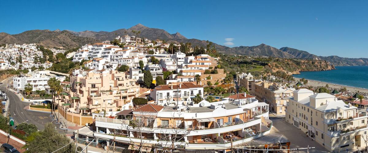 A coastal town with white buildings, a beach, and mountains in the background.