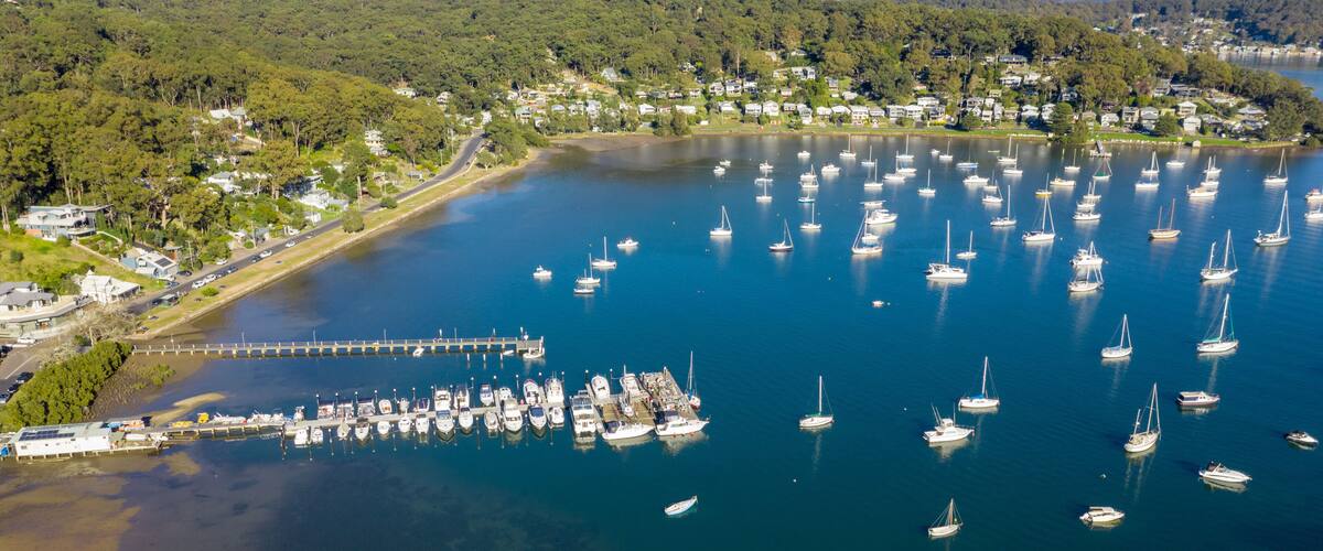 A marina with numerous sailboats docked, surrounded by a residential area and dense forest.